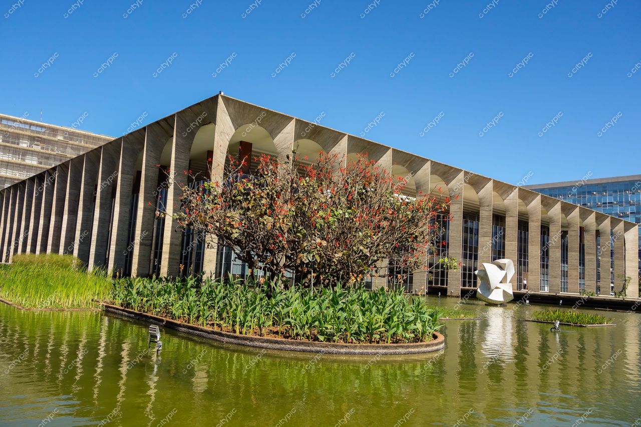 Palácio do Itamaraty em Brasília com lago, vegetação ao redor e escultura branca sob céu azul claro