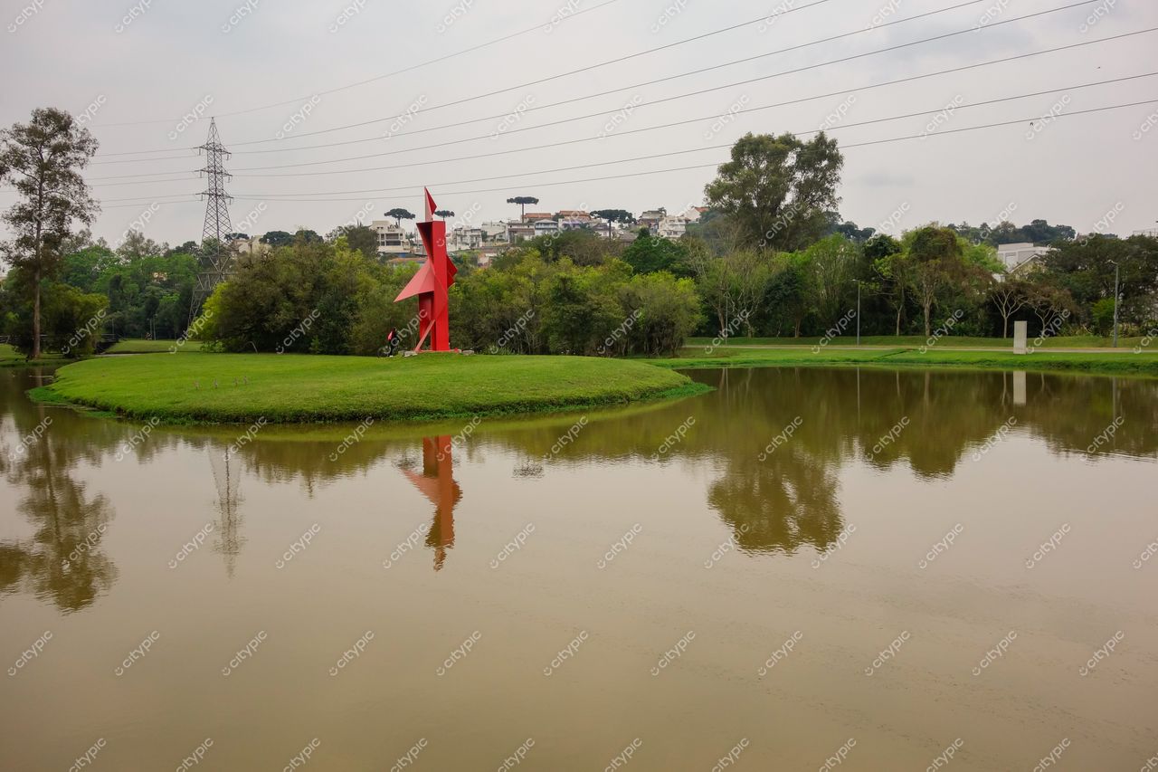 Parque Tingui em Curitiba com escultura vermelha refletida em lago sob céu nublado e vegetação densa