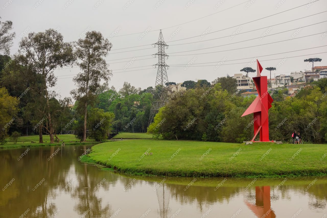 Parque Tingui em Curitiba com escultura vermelha angular, lago refletivo e vegetação densa sob céu nublado