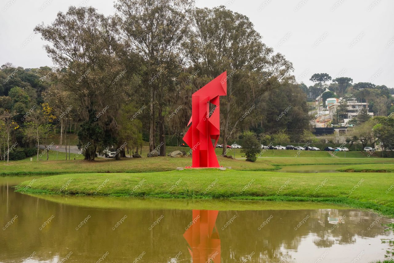 Parque Tingui em Curitiba com escultura moderna vermelha refletida no lago em dia nublado e vegetação ao fundo