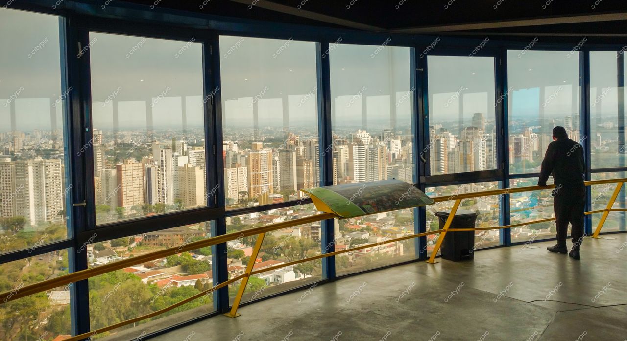 Torre Panorâmica de Curitiba em dia nublado com homem observando arranha-céus e áreas urbanas pela janela