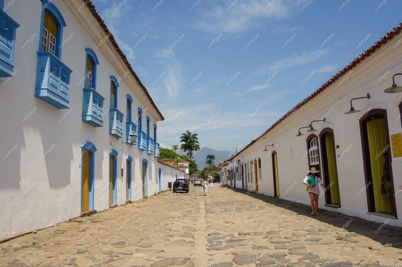 Paraty, Rio de Janeiro, rua histórica com casas coloniais brancas e detalhes em azul sob céu ensolarado e vegetação