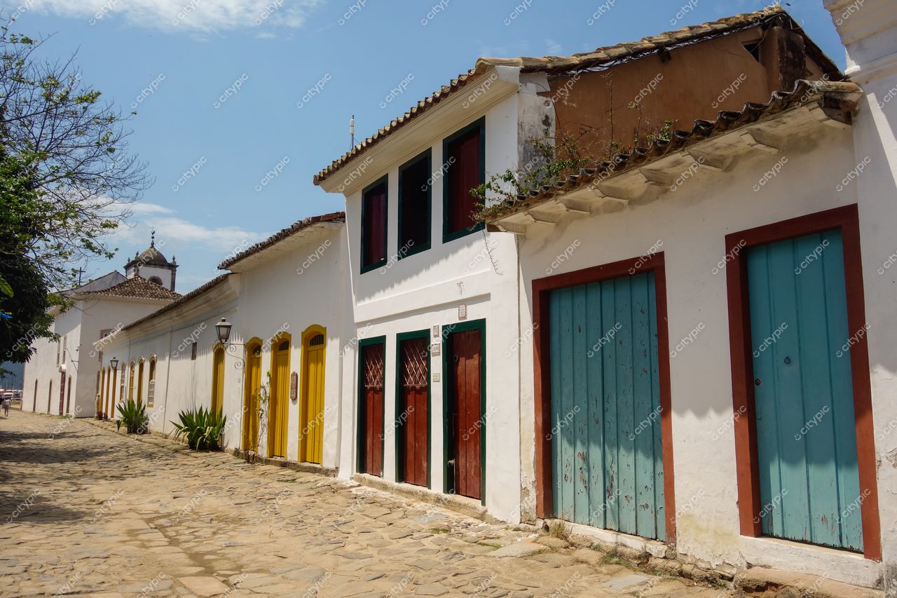 Paraty, Rio de Janeiro, rua tranquila com casas coloniais brancas e portas coloridas sob céu ensolarado e vegetação local