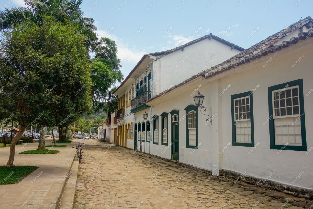 Paraty, Rio de Janeiro, rua tranquila com fachadas brancas e verdes, iluminação clara e vegetação ao longo da calçada