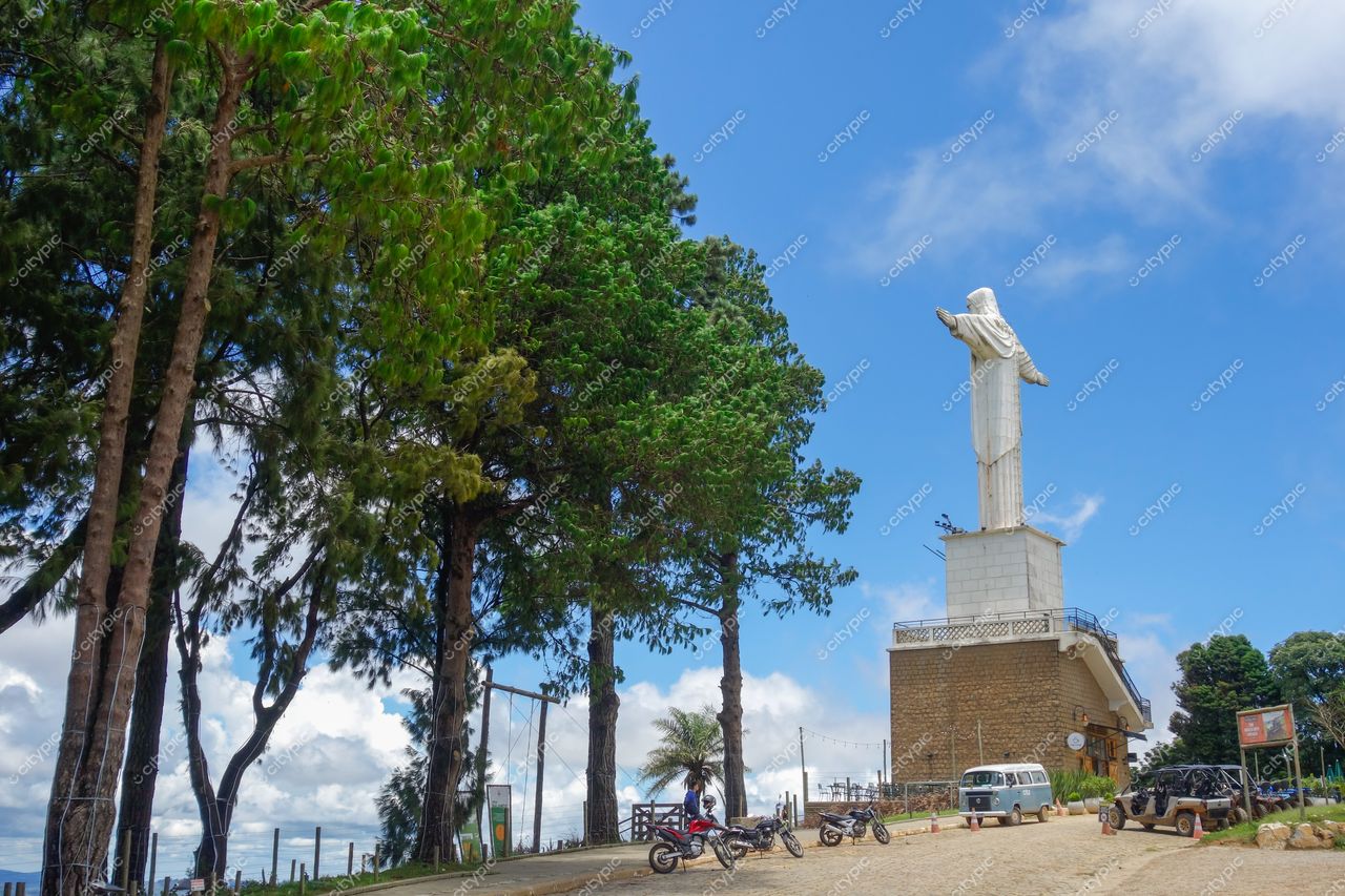 Cristo Redentor em Poços de Caldas com árvores altas, céu claro e veículos estacionados na estrada de terra