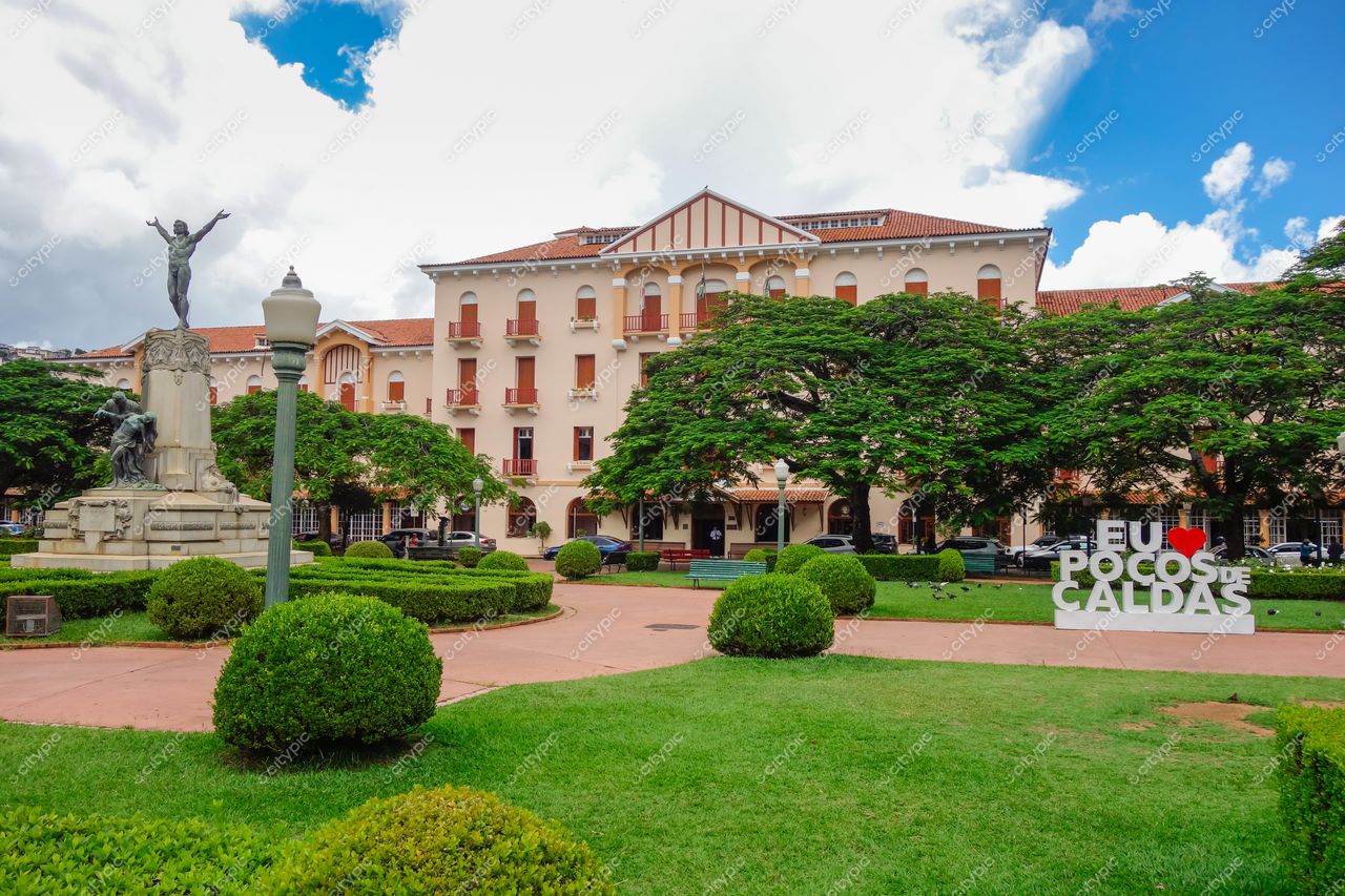 Praça Pedro Sanches e Palace Hotel em Poços de Caldas com estátua, vegetação exuberante e céu parcialmente nublado