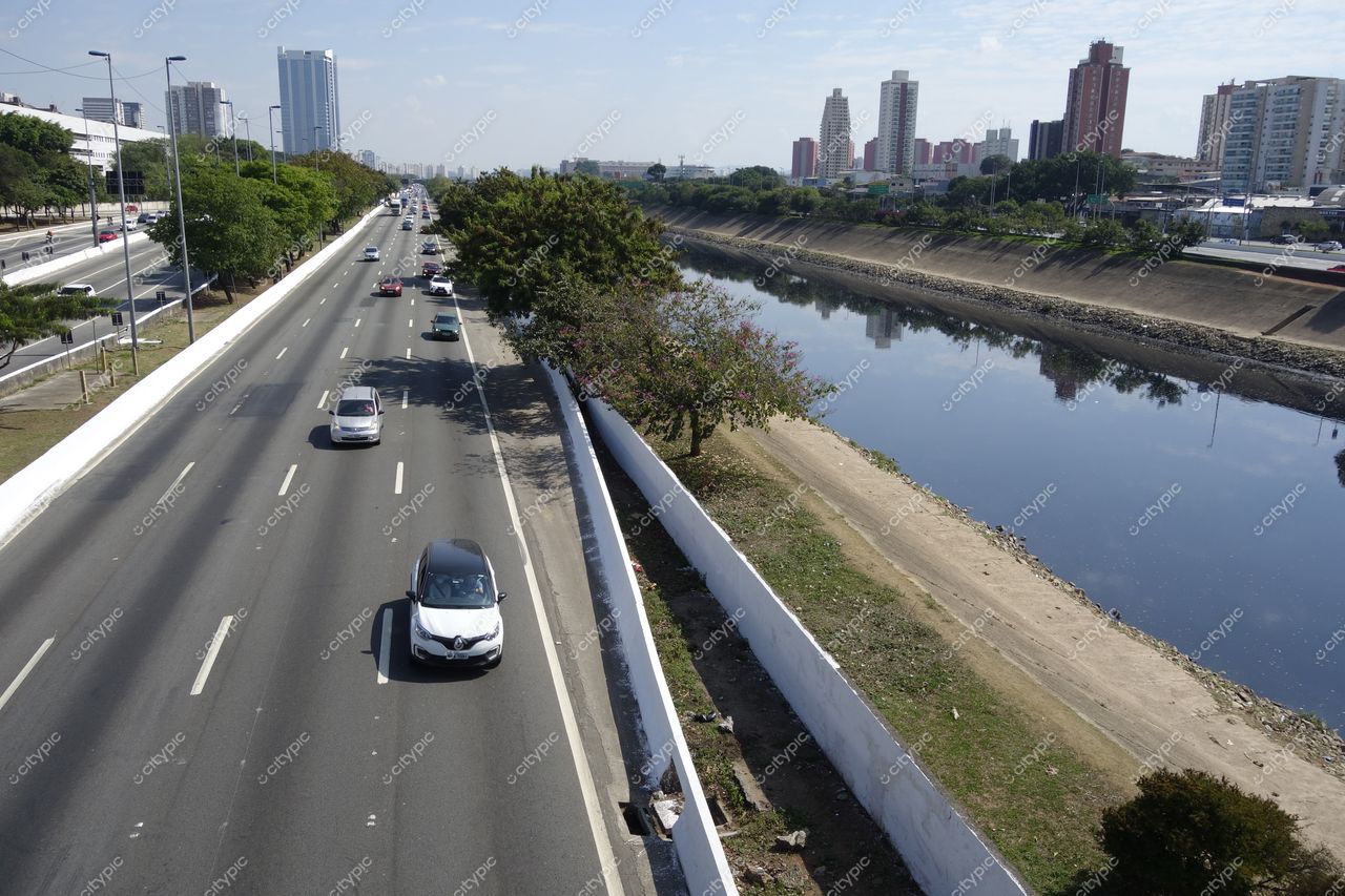 São Paulo, São Paulo, via movimentada com carros, árvores à esquerda e rio calmo à direita sob céu ensolarado