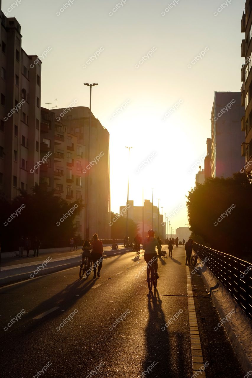 São Paulo, São Paulo, pôr do sol com ciclistas e pedestres em via urbana com vegetação e silhuetas de edifícios