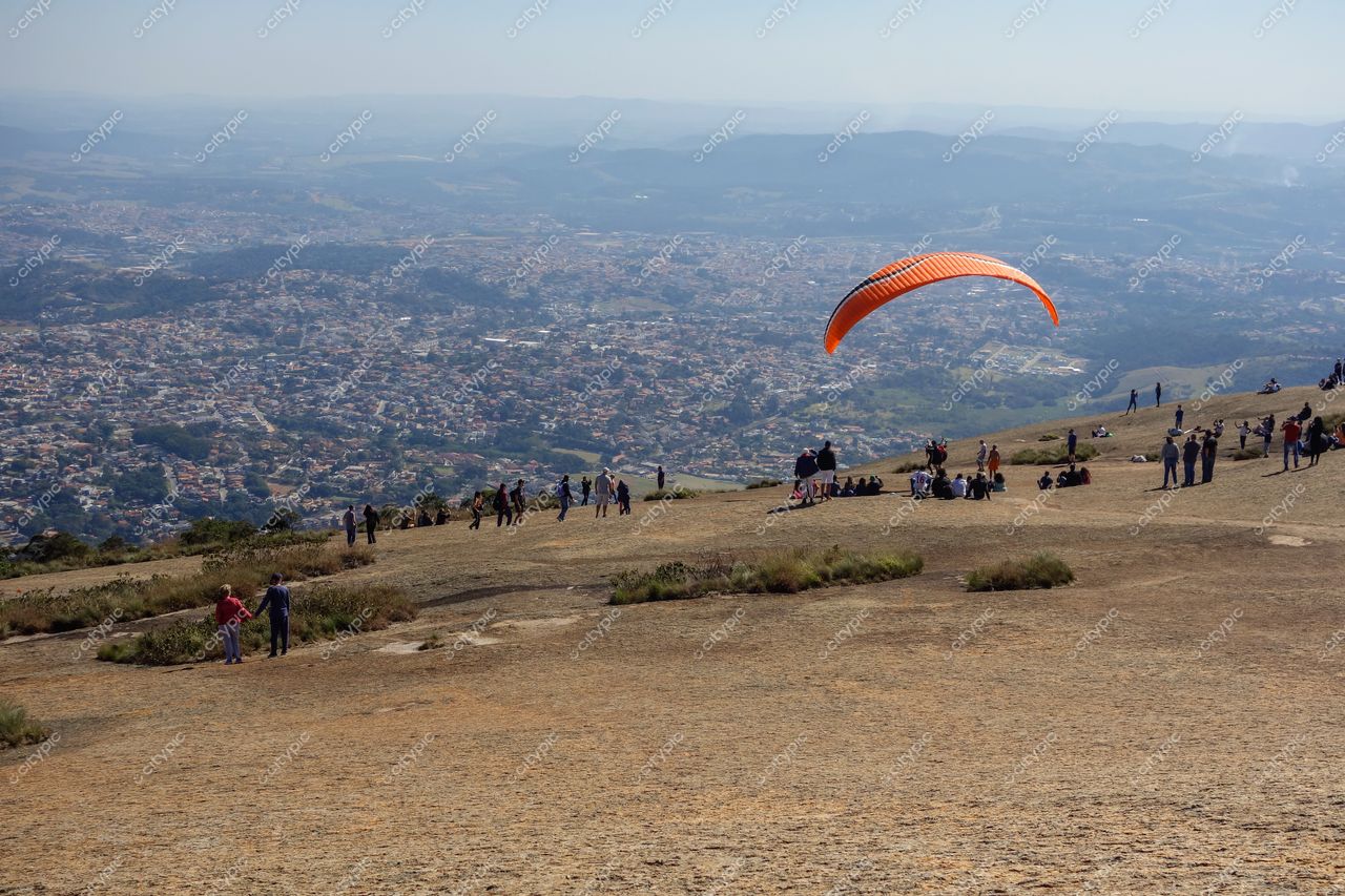 Atibaia, São Paulo, vista panorâmica em dia ensolarado com parapente laranja e campo rochoso repleto de pessoas