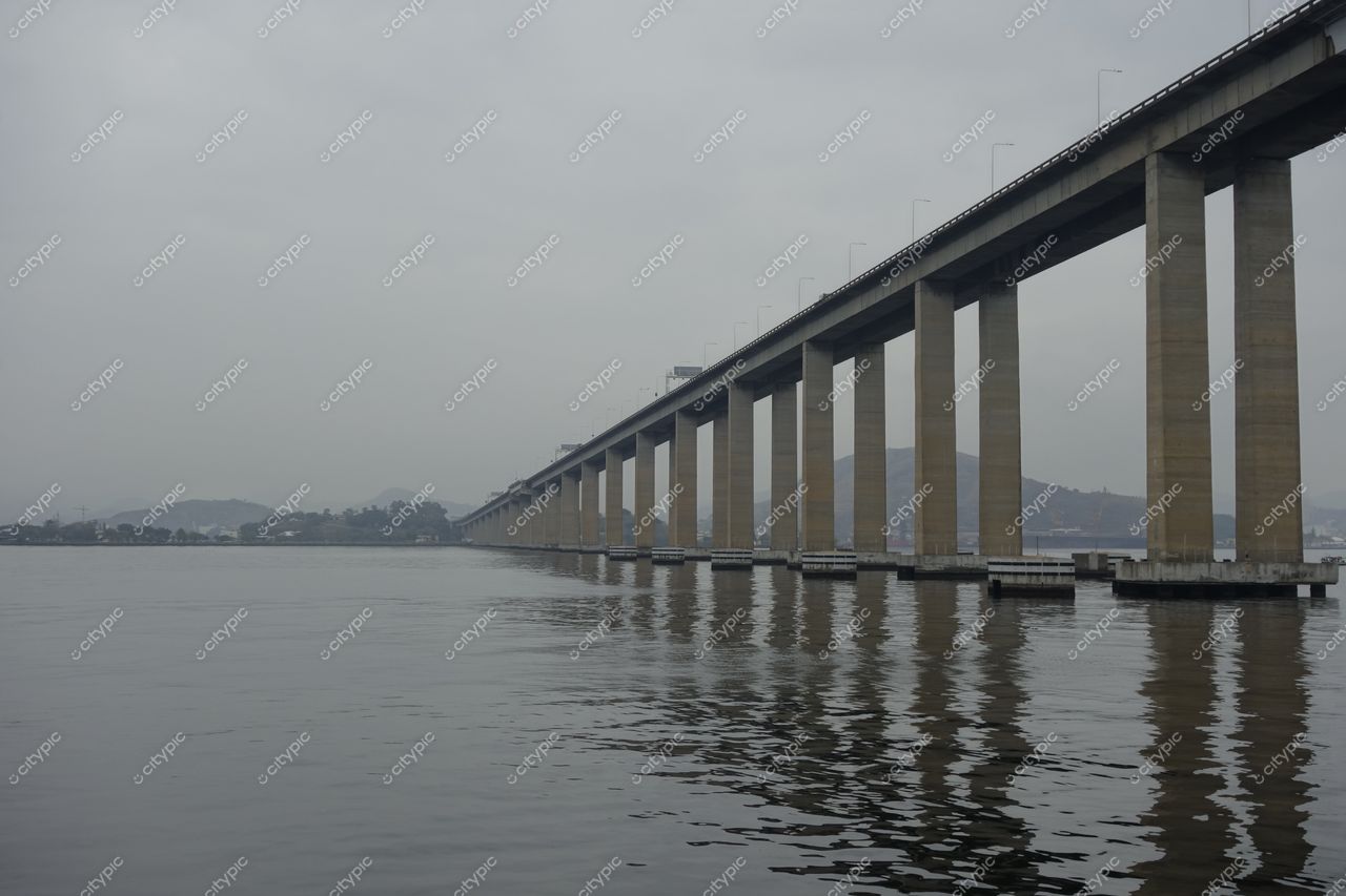 Rio de Janeiro, Rio de Janeiro, ponte elevada refletindo na água calma sob céu nublado e montanhas ao fundo