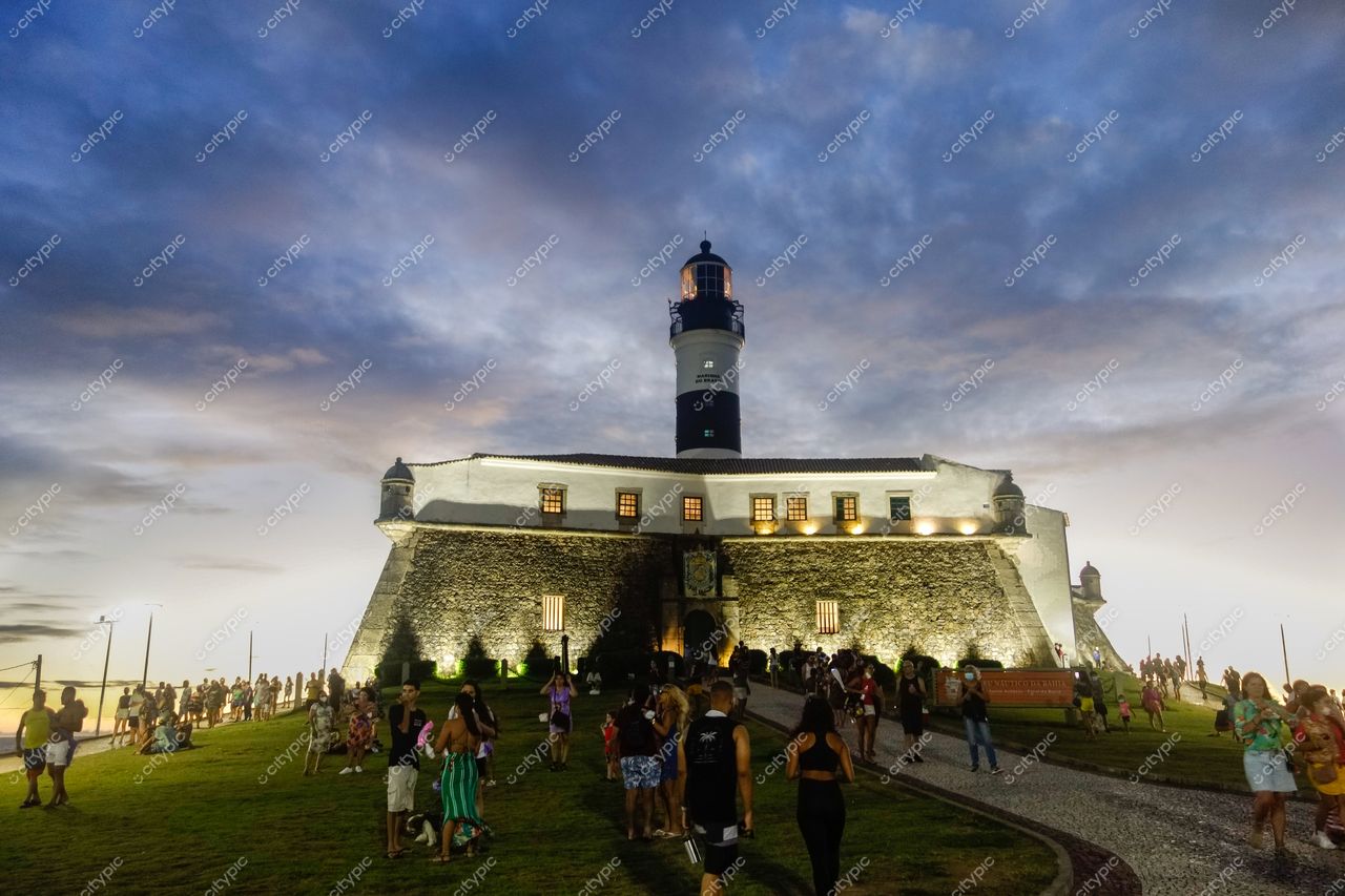Farol da Barra em Salvador ao crepúsculo com céu azul e rosa, pessoas reunidas no gramado e iluminação suave
