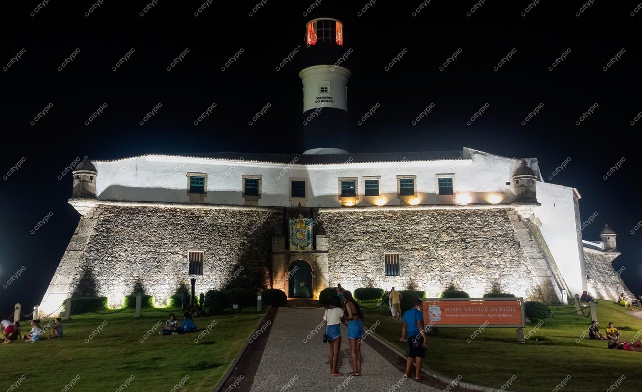Farol da Barra em Salvador iluminado à noite com pessoas reunidas e placa do Museu Náutico da Bahia ao fundo
