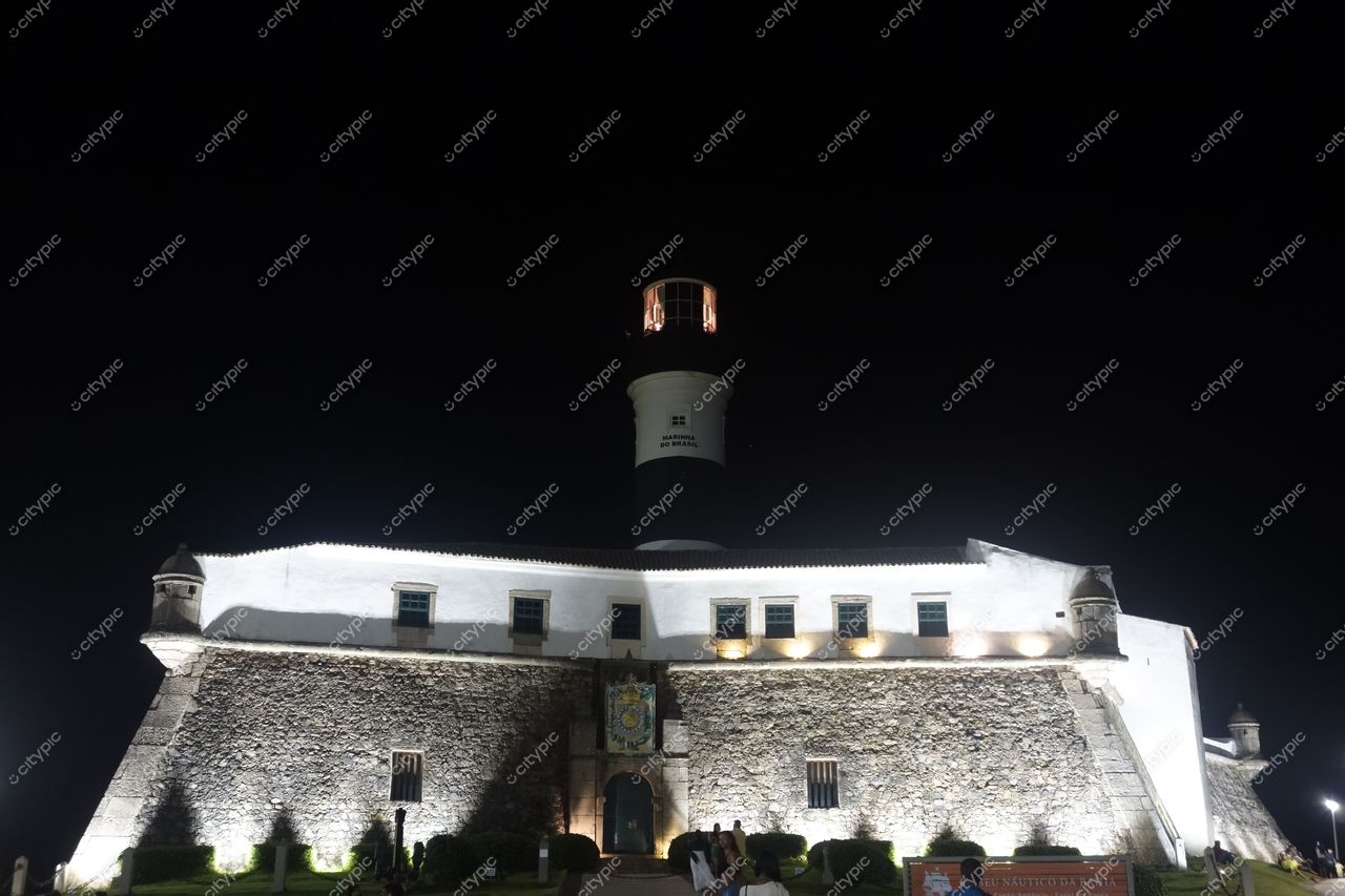 Farol da Barra em Salvador iluminado à noite com pessoas ao redor e vegetação à beira-mar em destaque