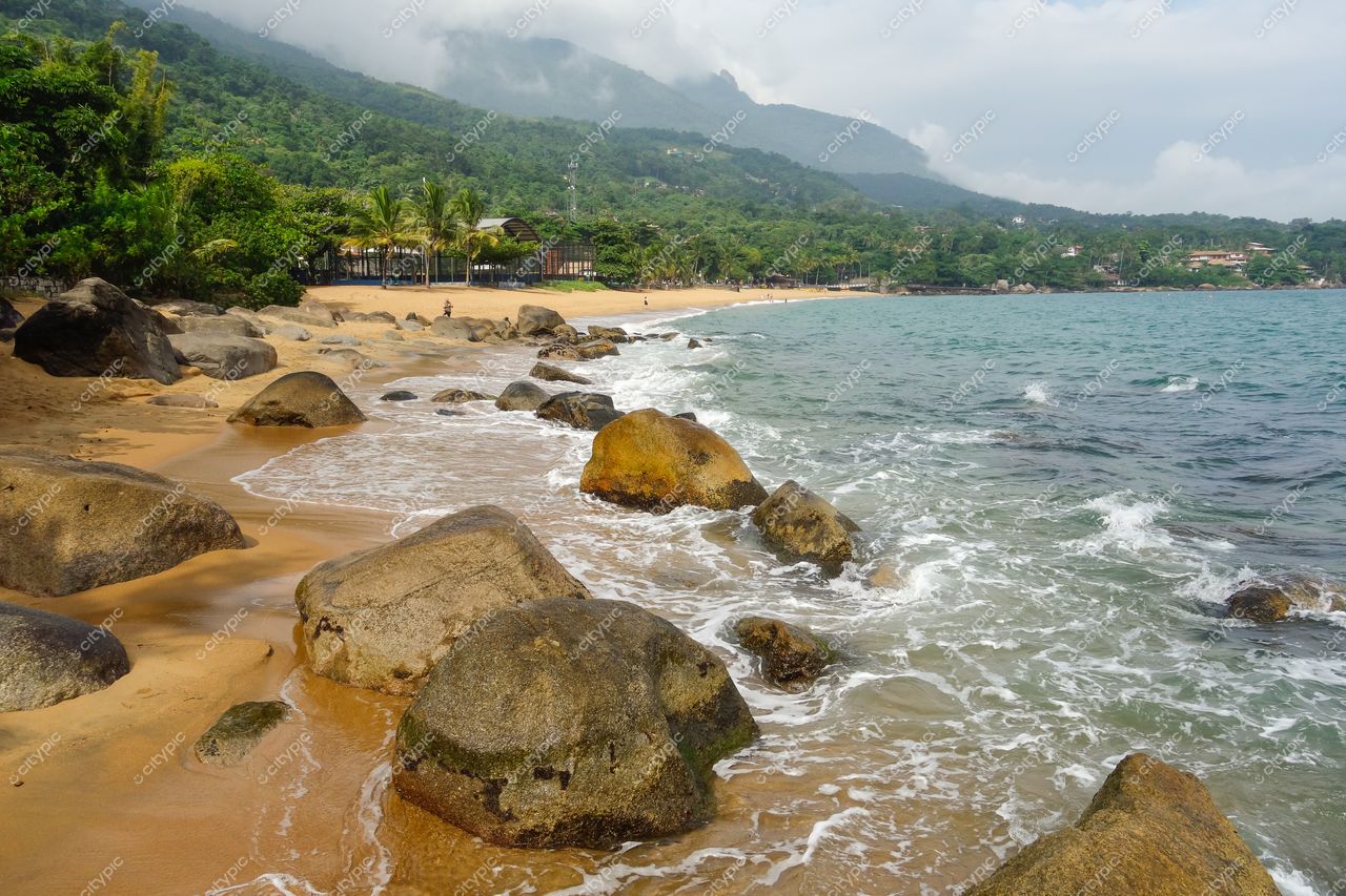 Ilhabela, São Paulo, praia com areia dourada, grandes pedras cobertas de musgo e montanhas arborizadas ao fundo