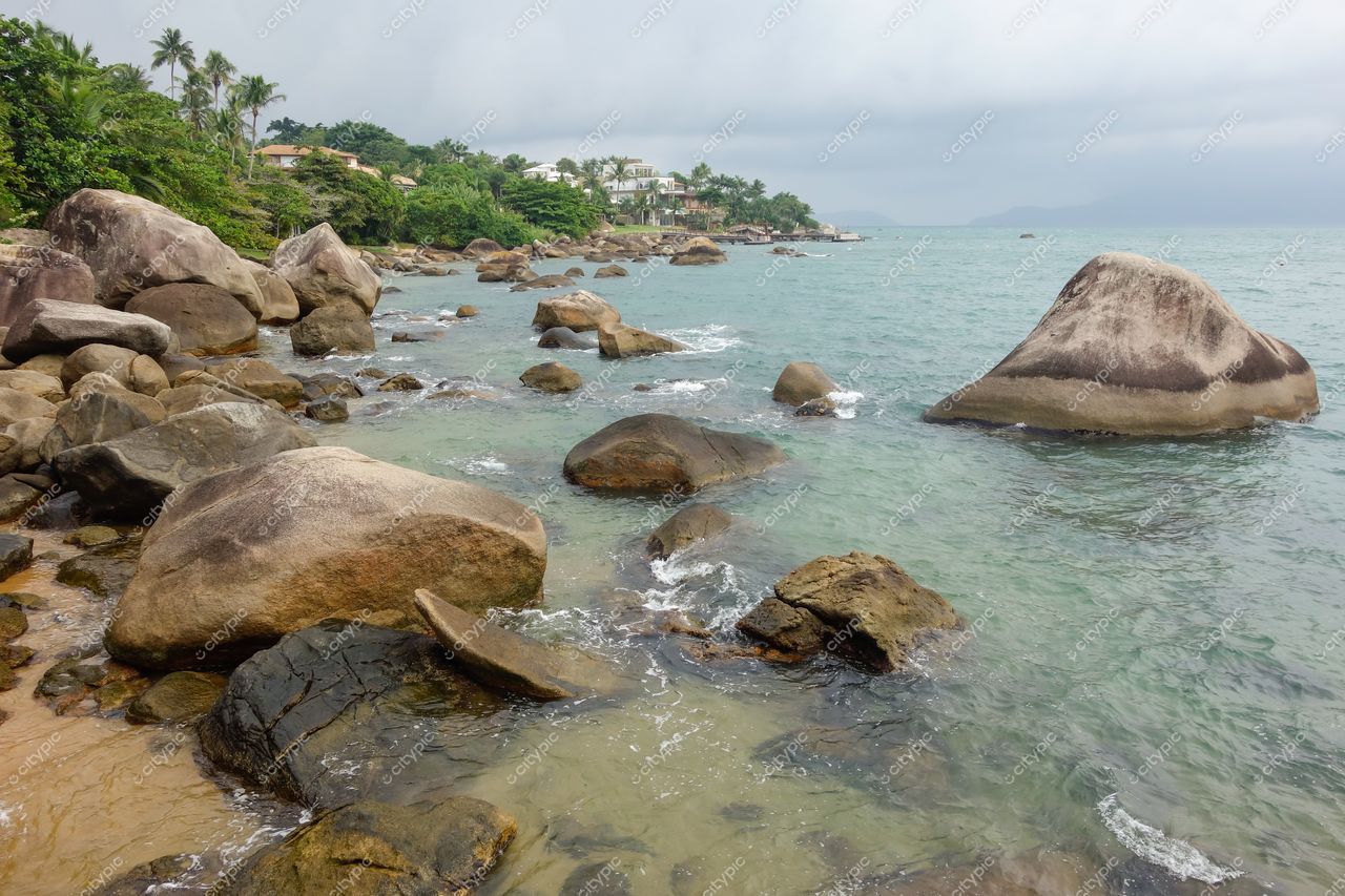 Ilhabela, São Paulo, costa rochosa com grandes pedras e águas verdes em dia nublado com vegetação densa ao fundo