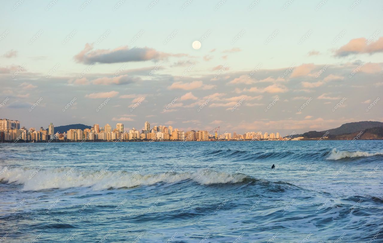 Santos, São Paulo, vista do mar ao amanhecer com lua cheia e edifícios refletindo a luz suave da manhã