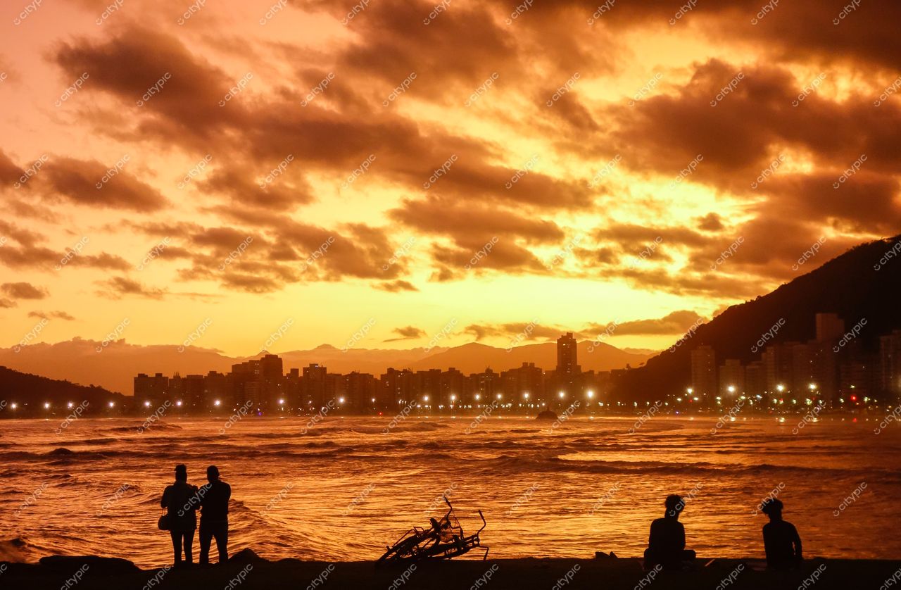 Santos, São Paulo, pôr do sol na praia com nuvens alaranjadas e silhuetas de pessoas observando o mar agitado
