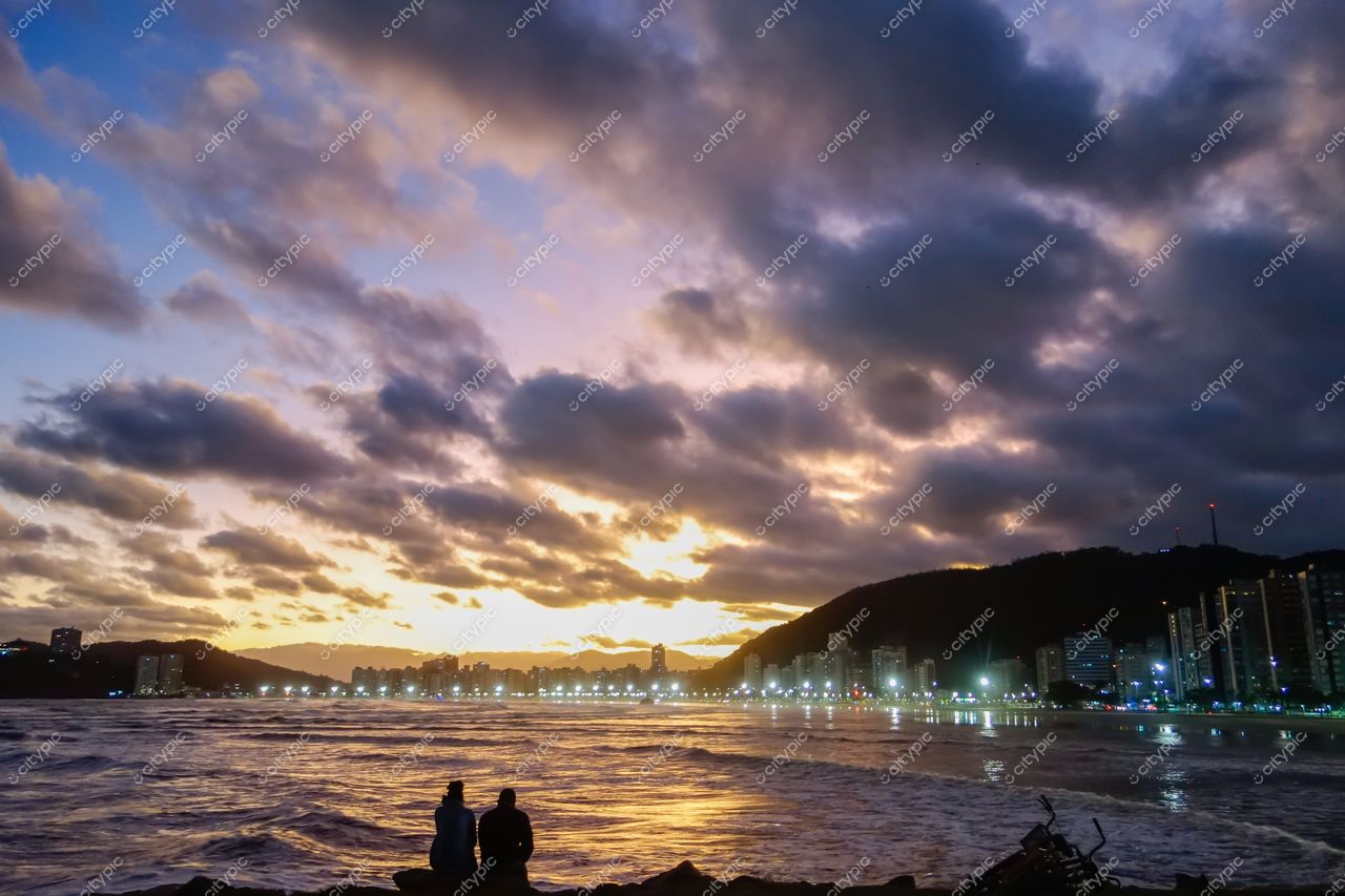 Santos, São Paulo, pôr do sol na praia com casal em rochas e edifícios iluminados ao fundo