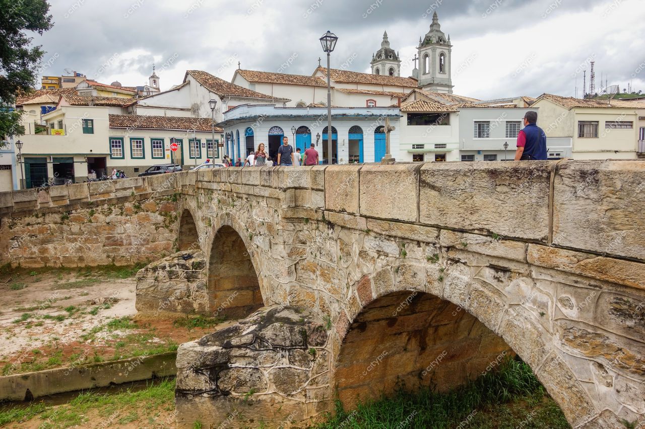 Ponte da Cadeia em São João del Rei com arcos de pedra e céu nublado, destacando a vegetação e o tráfego de pedestres