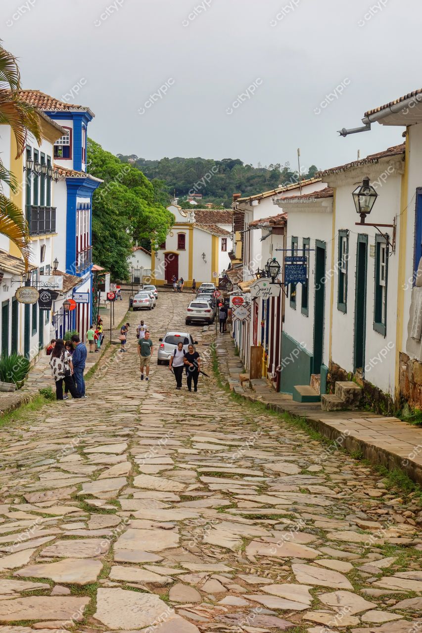 Tiradentes, Minas Gerais, rua de paralelepípedos com casas coloniais em dia nublado e vegetação ao fundo