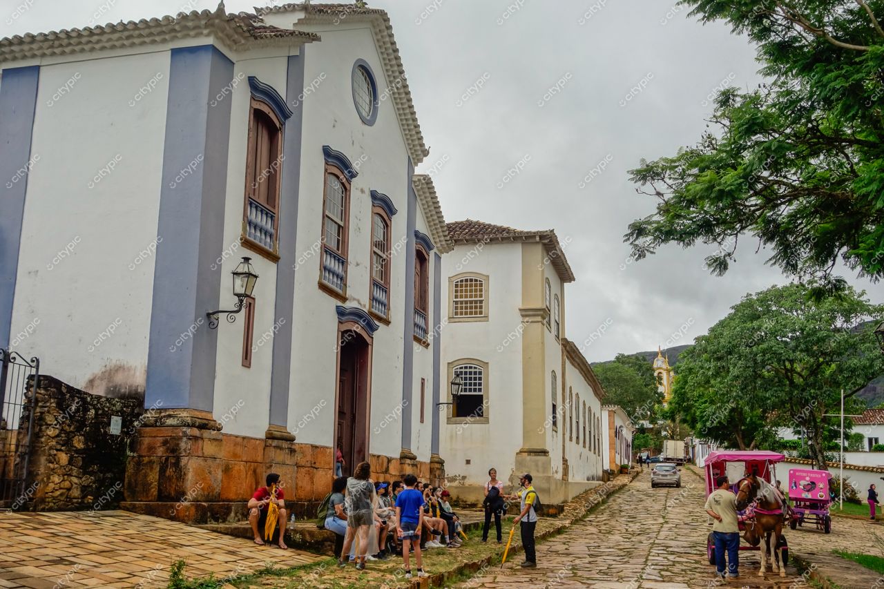 Tiradentes, Minas Gerais, rua histórica com arquitetura colonial, céu nublado e vegetação ao redor, vida cotidiana e turismo