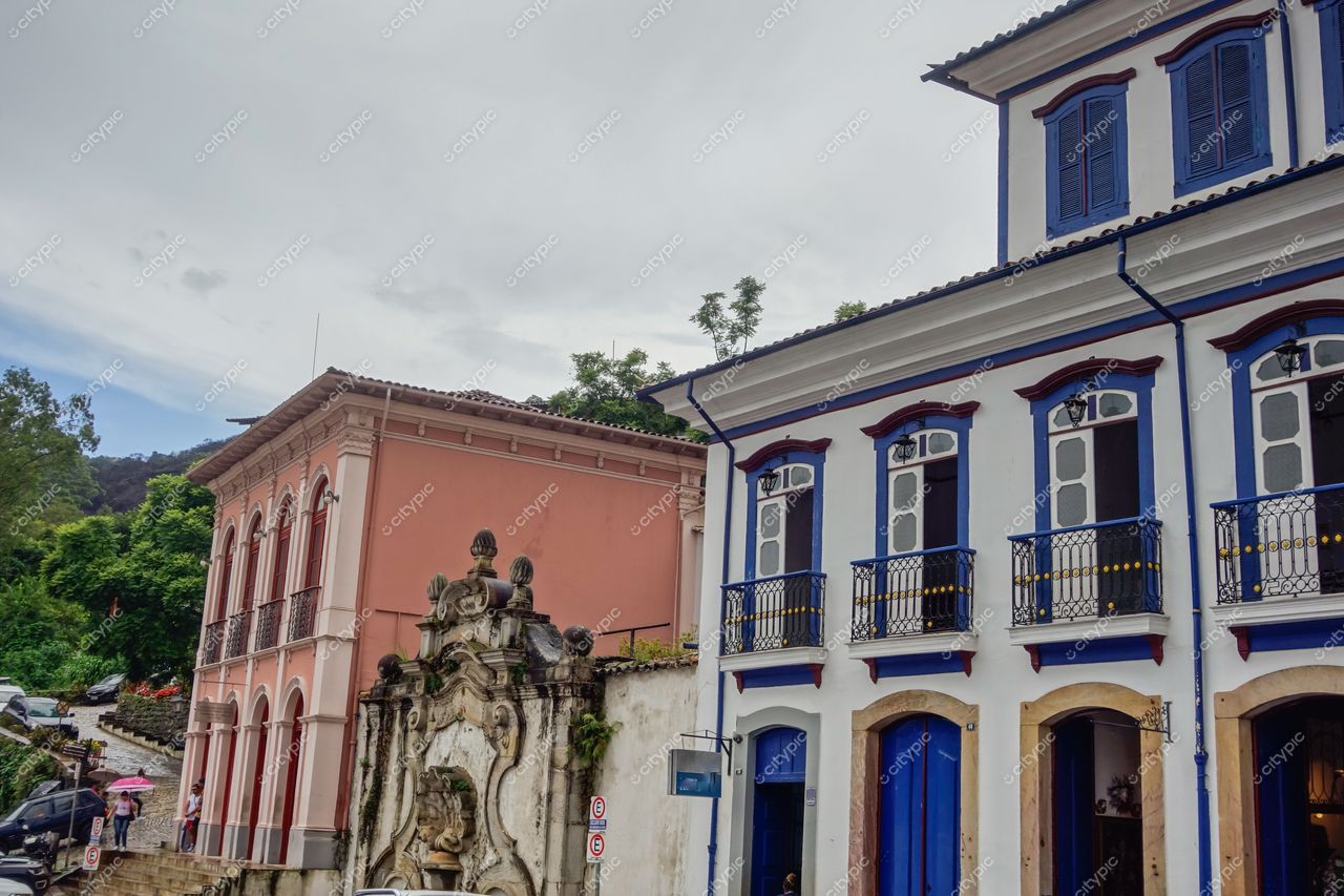 Ouro Preto, Minas Gerais, edifícios históricos rosa e branco sob céu nublado com fonte ornamentada e vegetação verde