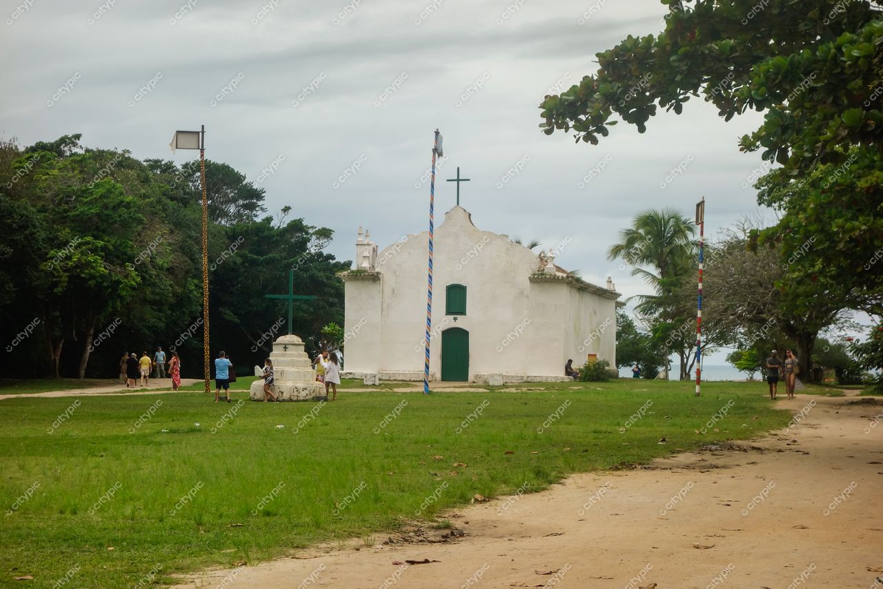 Igreja Matriz Nossa Senhora d’Ajuda em Arraial d’Ajuda com céu nublado e pessoas caminhando pelo caminho de terra