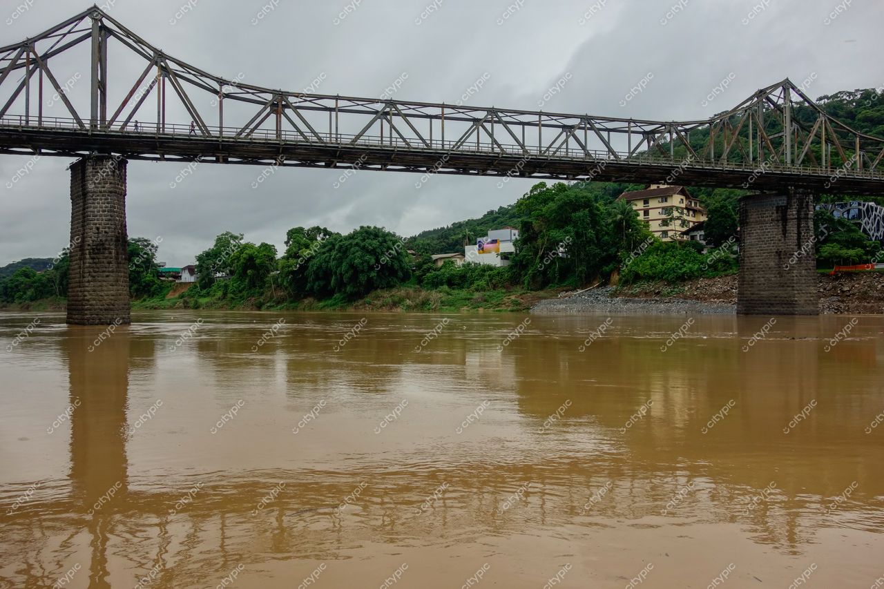Blumenau, Santa Catarina, ponte metálica sobre rio marronada em dia nublado com vegetação densa e edificações urbanas