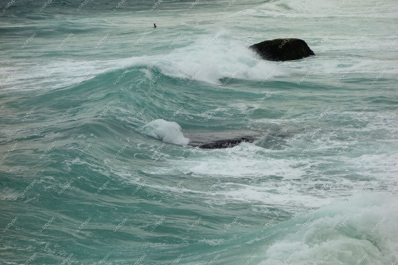 Florianópolis, Santa Catarina, mar agitado com ondas em azul e verde, rochedo visível e nadador em dia nublado