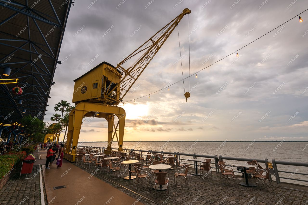 Belém, Pará vista à beira da água com guindaste amarelo ao entardecer e mesas na calçada junto ao rio