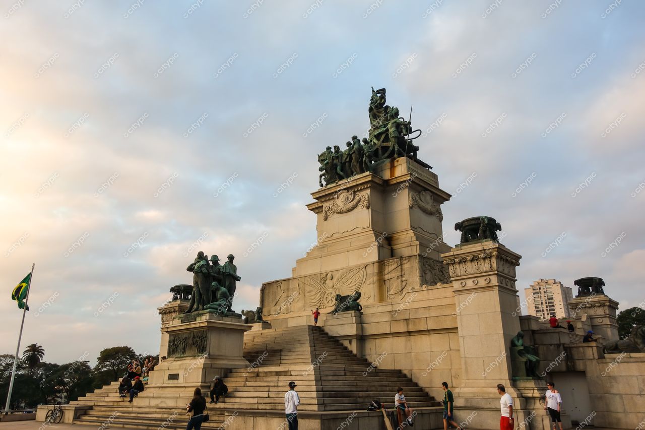 Monumento às Bandeiras em São Paulo com céu nublado e iluminação suave ao entardecer, rodeado por vegetação e visitantes