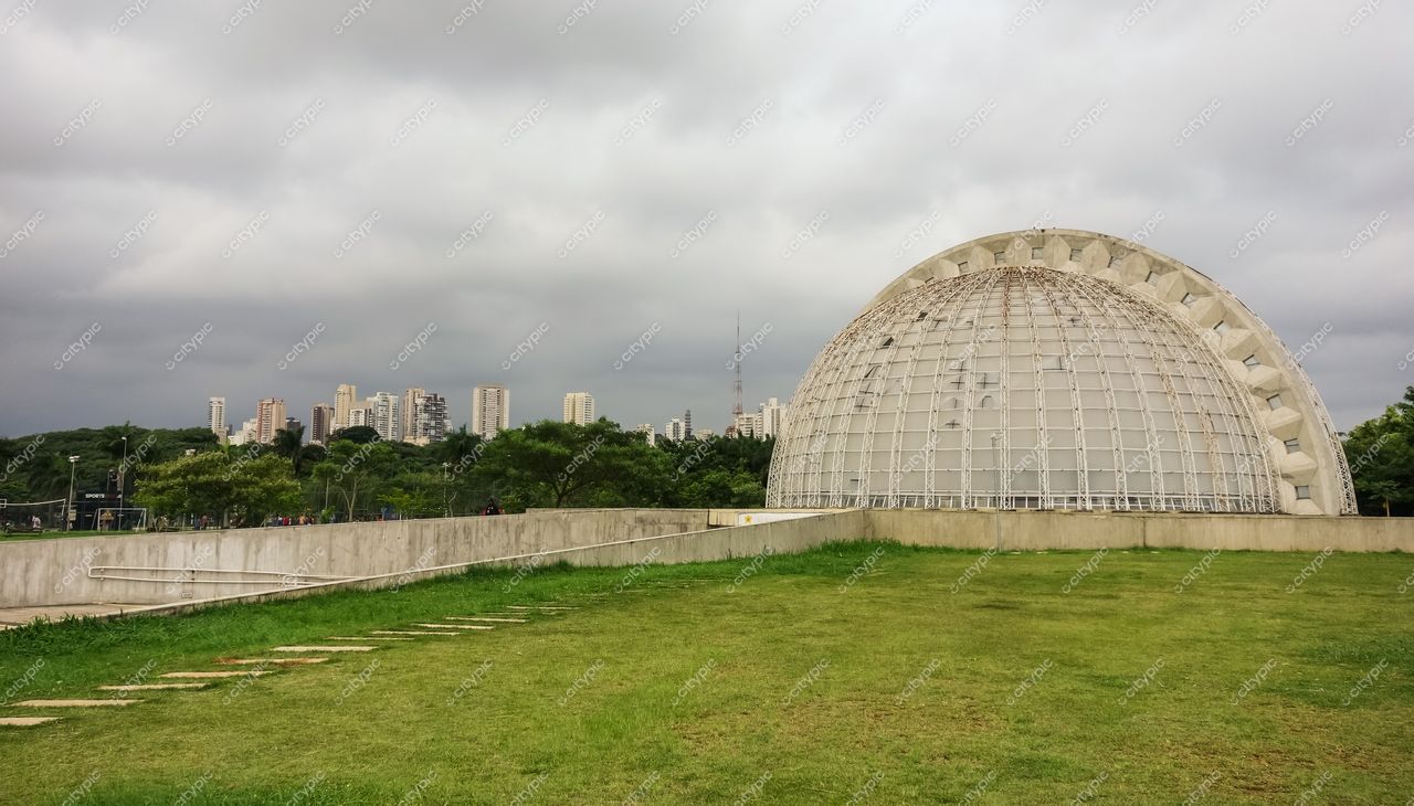 Parque Ibirapuera, em São Paulo, com edifício moderno semicircular sob céu nublado e horizonte urbano ao fundo