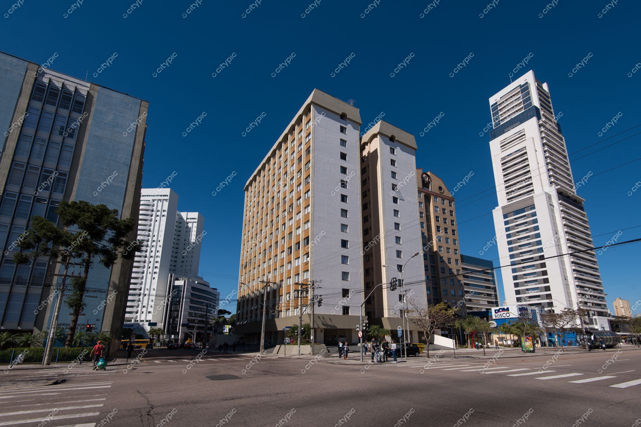 Curitiba, Paraná, interseção movimentada com arquitetura moderna e árvores alinhadas sob céu azul intenso durante o dia