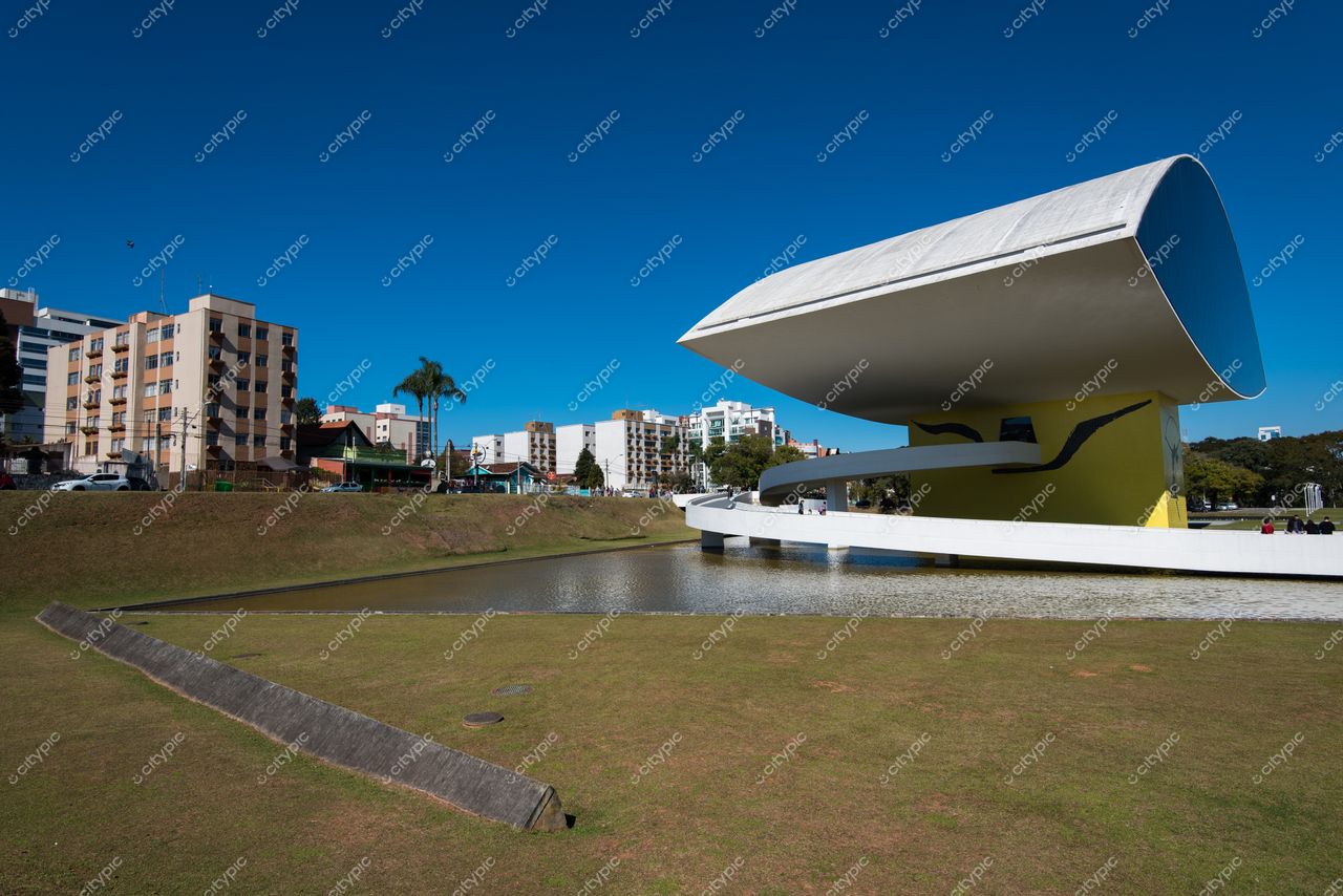 Curitiba, Paraná, edifício moderno com cobertura curva à beira de lago em dia ensolarado e áreas verdes