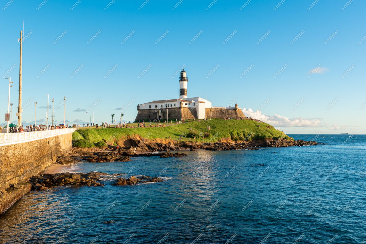 Salvador, Bahia, Farol da Barra em dia ensolarado com pessoas caminhando na orla e vegetação ao redor