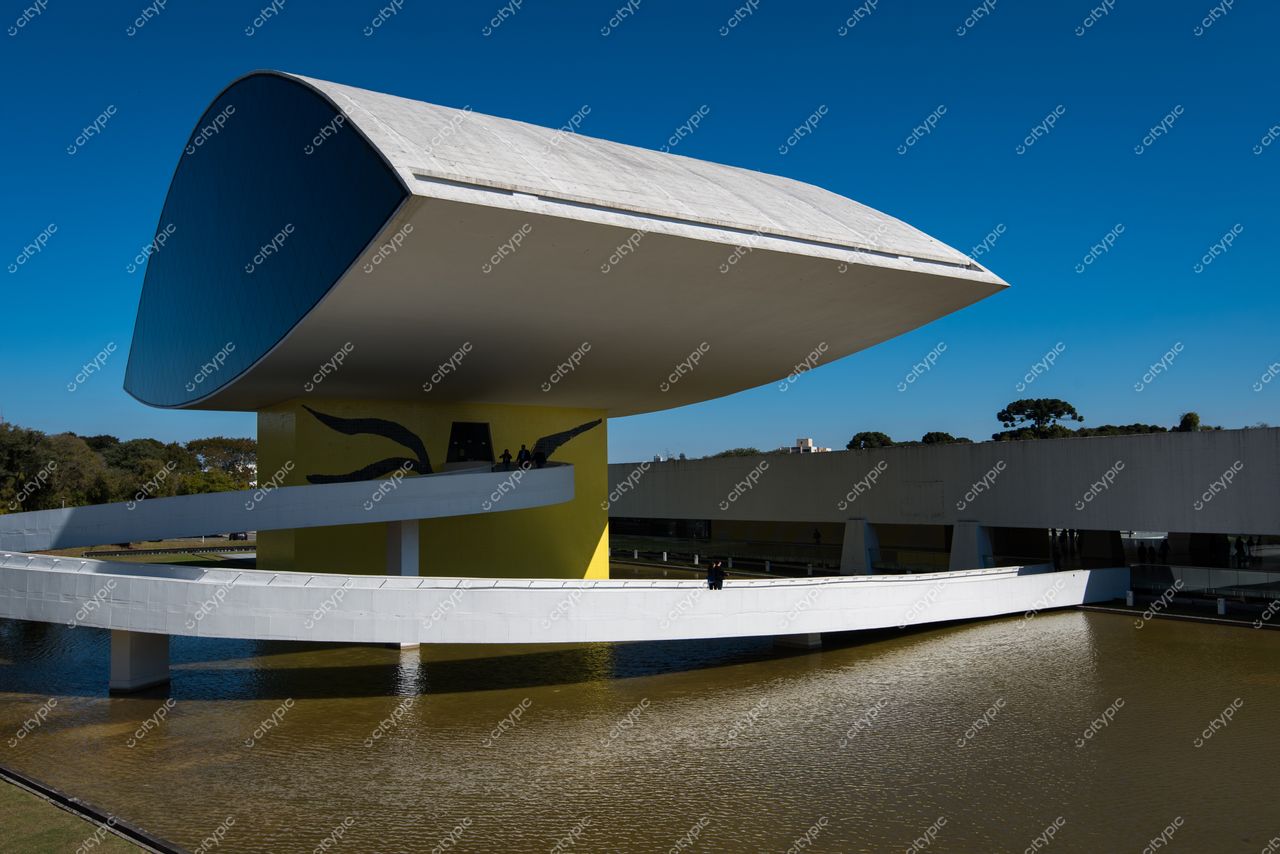 Curitiba, Paraná, edifício moderno com design arqueado refletindo luz do sol em dia claro e vegetação ao fundo