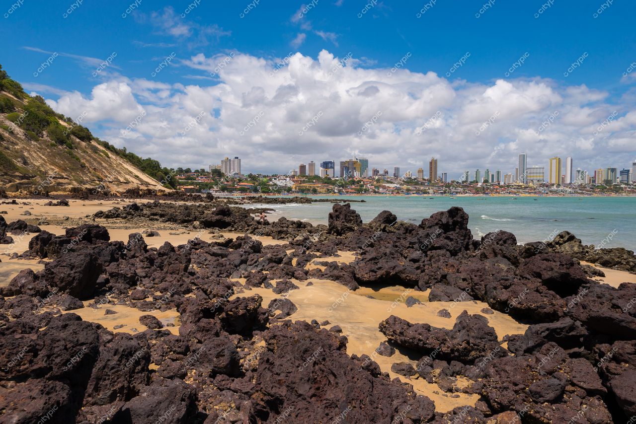 Natal, Rio Grande do Norte com mar esverdeado e céu nublado, rochas na areia e perfil urbano ao fundo