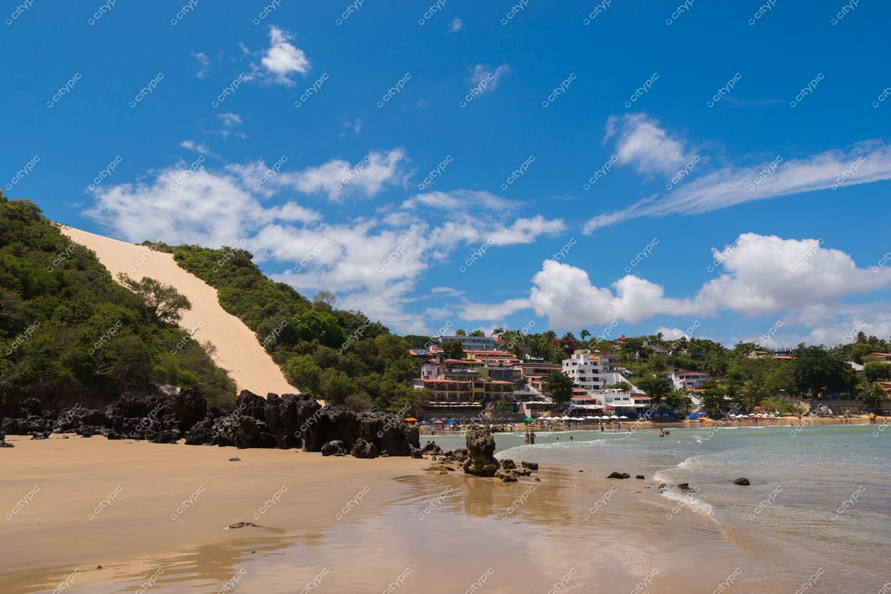 Natal, Rio Grande do Norte, praia com areia clara, morro verde ao fundo e casas em destaque sob céu azul