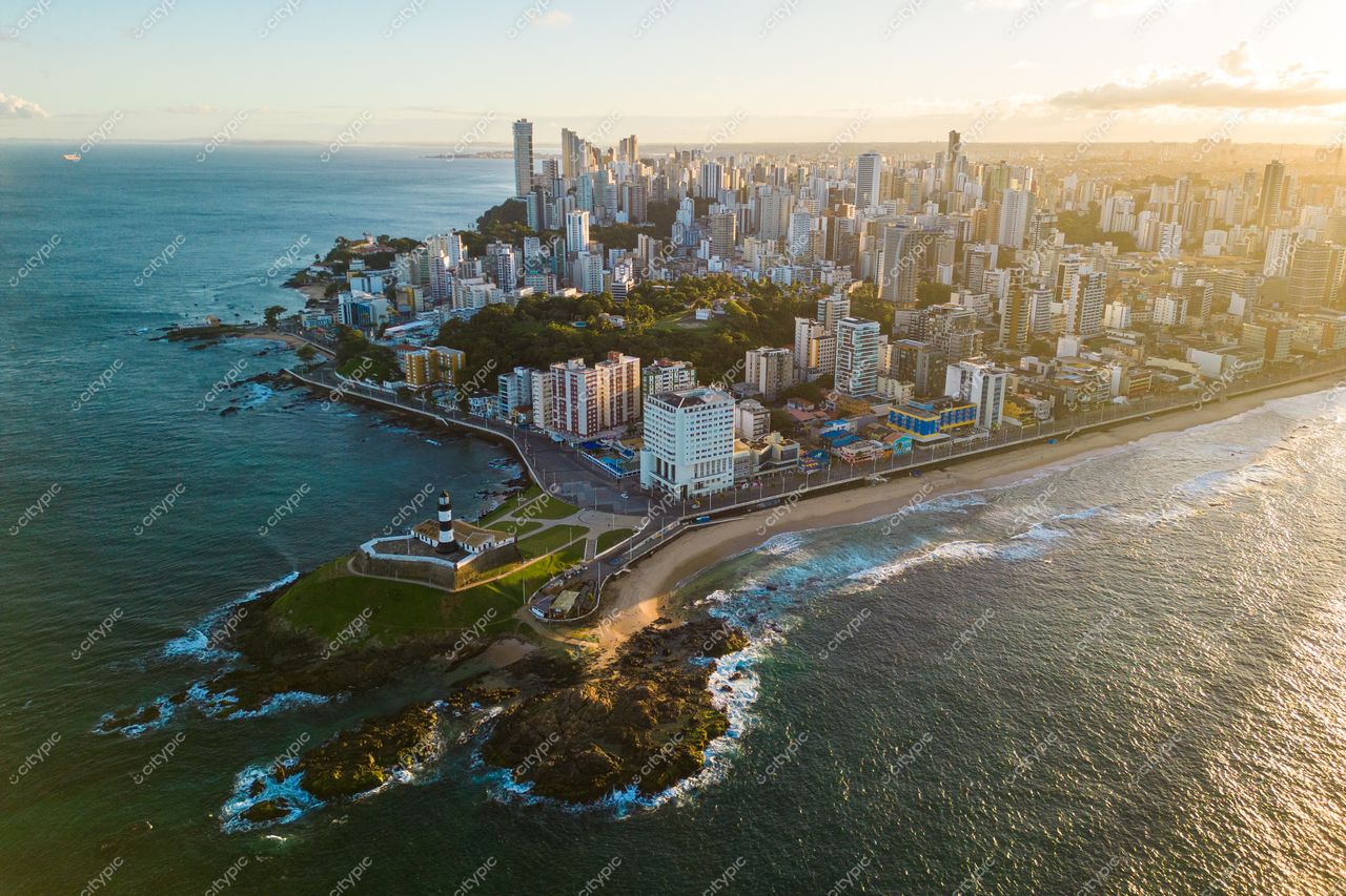 Salvador, Bahia vista aérea com costa à esquerda, skyline urbano moderno e farol em morro vegetado ao entardecer