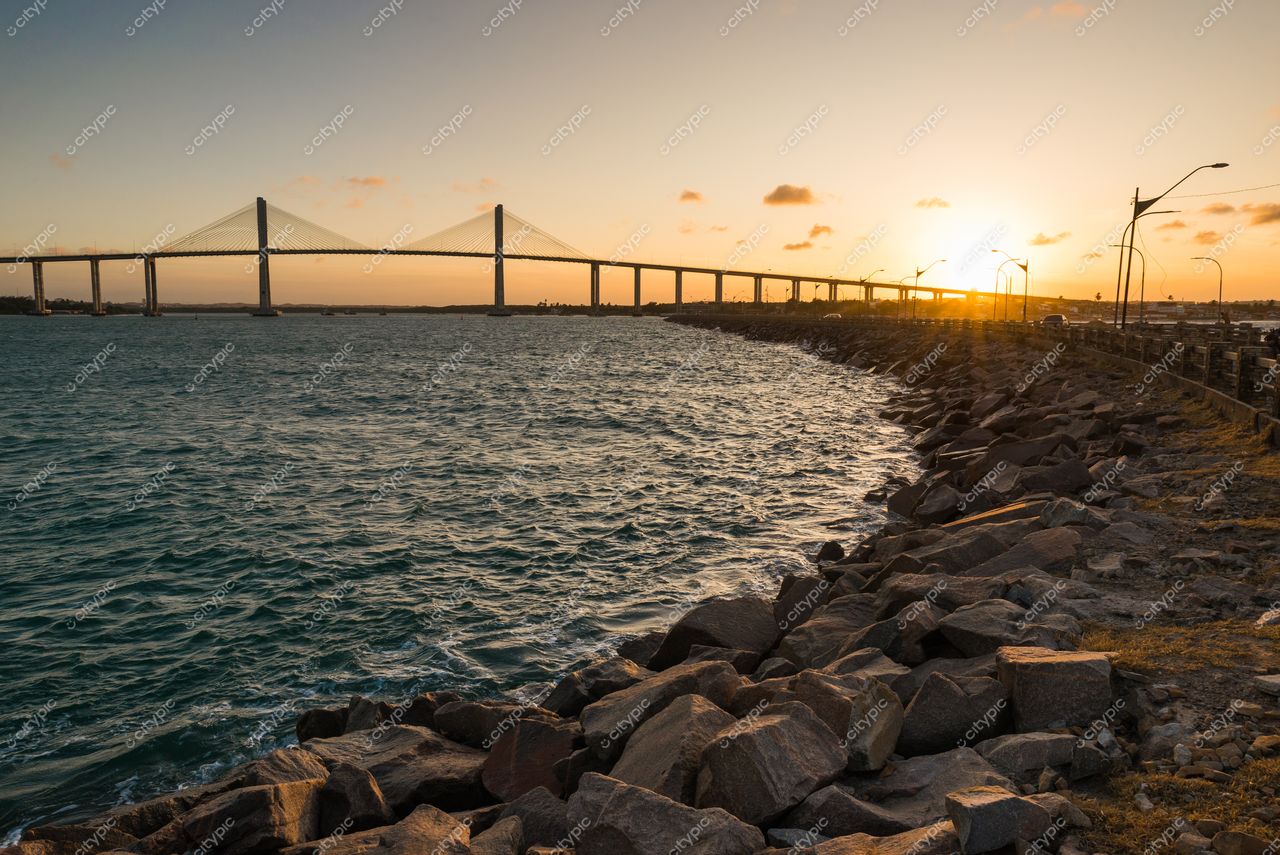 Natal, Rio Grande do Norte, pôr do sol com ponte suspensa refletindo luz na baía e pedras na costa