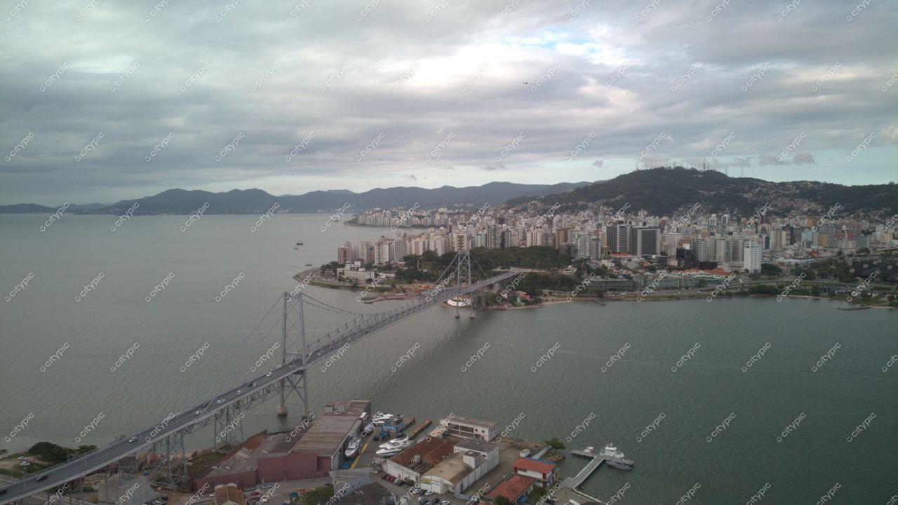 Centro de Florianópolis com vista panorâmica da ponte, arranha-céus e montanhas sob céu nublado e luz difusa