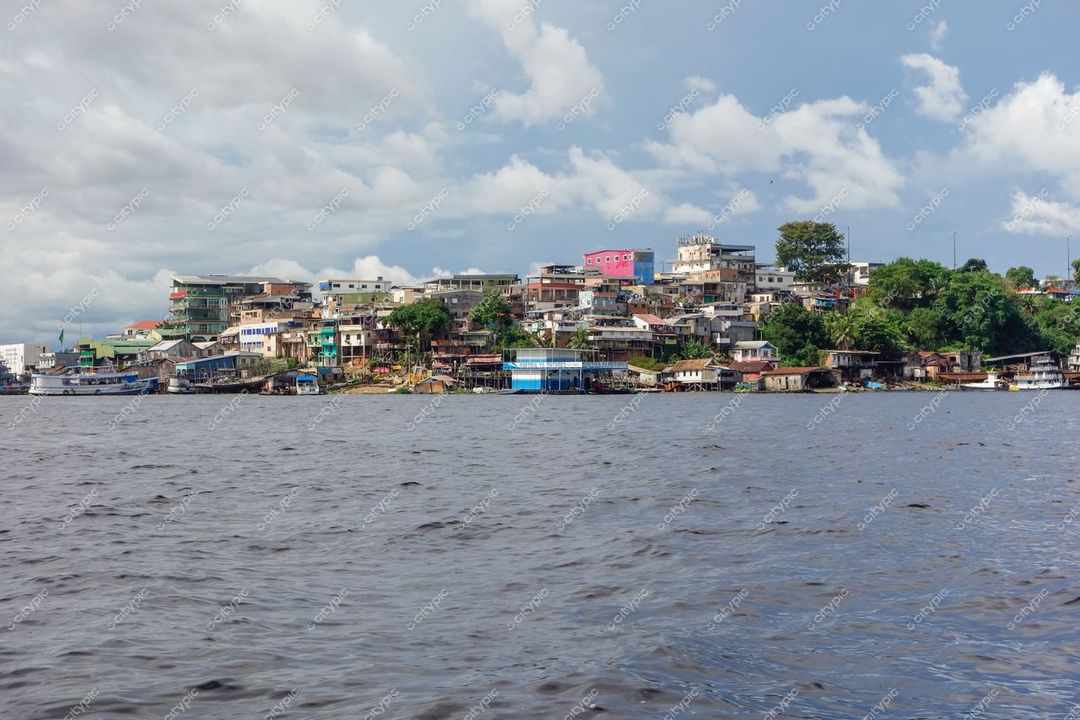 Manaus, Amazonas, vista do rio com casas coloridas em colina e vegetação ao redor sob céu parcialmente nublado