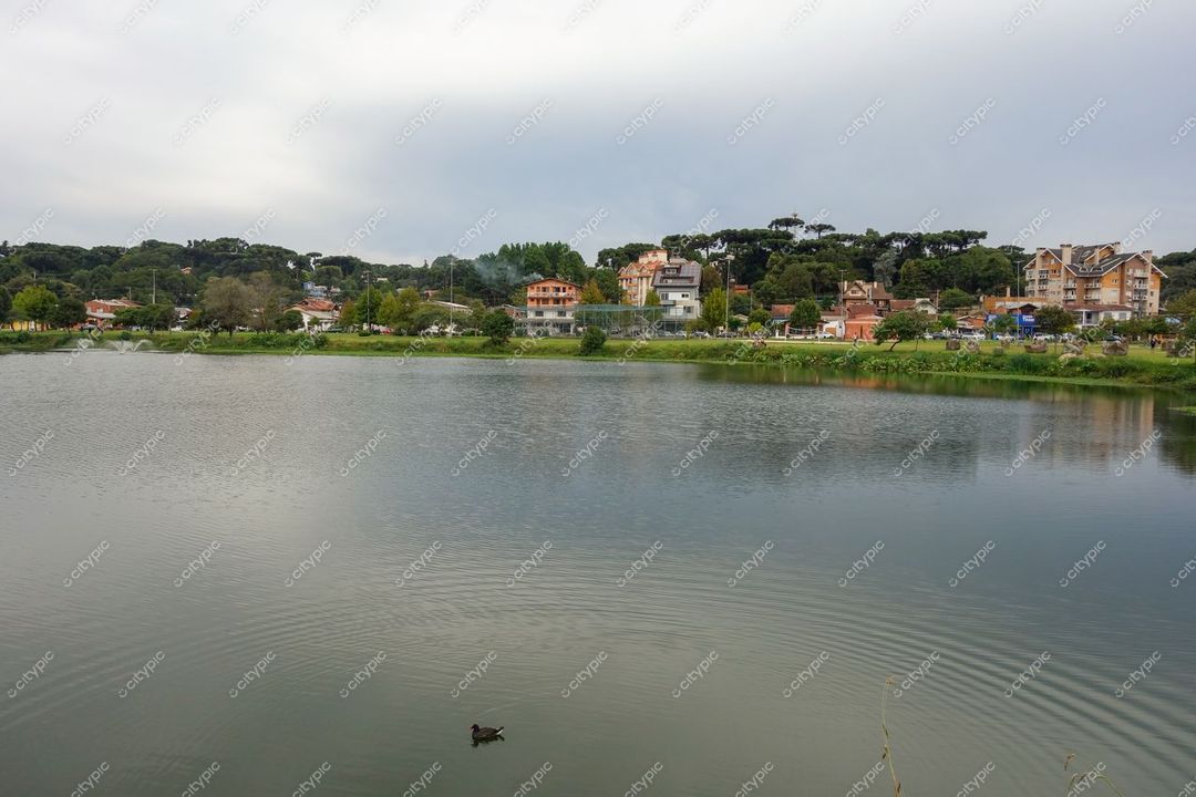 Canela, Rio Grande do Sul apresenta lago sereno com vegetação exuberante e casas refletidas sob céu nublado